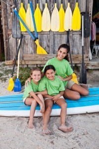 Teenage girl (17 years) with children (8-9 years) sitting on paddle board by water sports equipment center